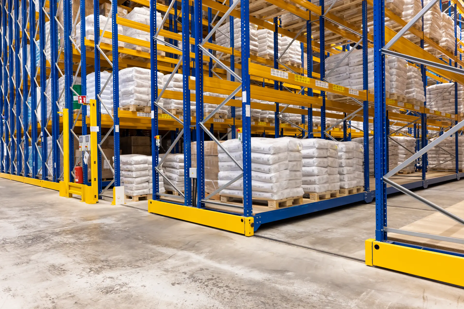 מדפי מנהלים ניידים בצבעי כחול־צהוב פועלים במערכת מסילות רצפתית, ואופני הפלטה מזיזים יחידות מדפים זו לצד זו